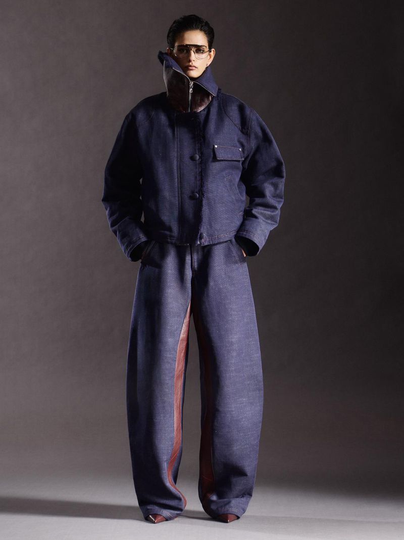 A slender figure poses in a striking blue denim jacket and baggy pants, accented by a unique brown leather panel and statement collar.