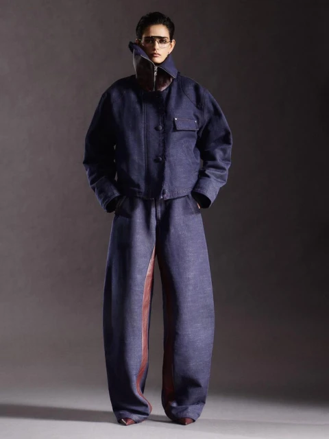 A slender figure poses in a striking blue denim jacket and baggy pants, accented by a unique brown leather panel and statement collar.