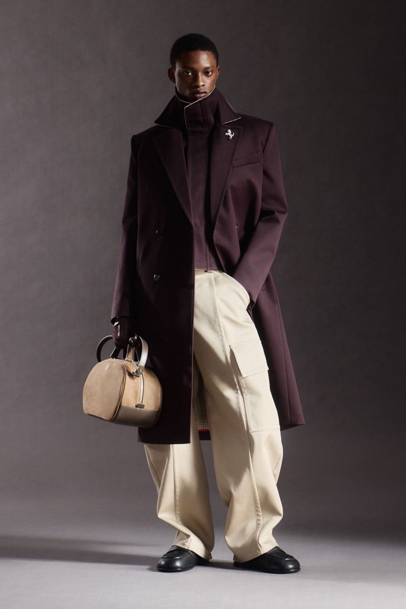 Model Nonso Ojukwu in a dark burgundy high-collar overcoat and beige wide-leg trousers holding a tan duffle bag in a studio.