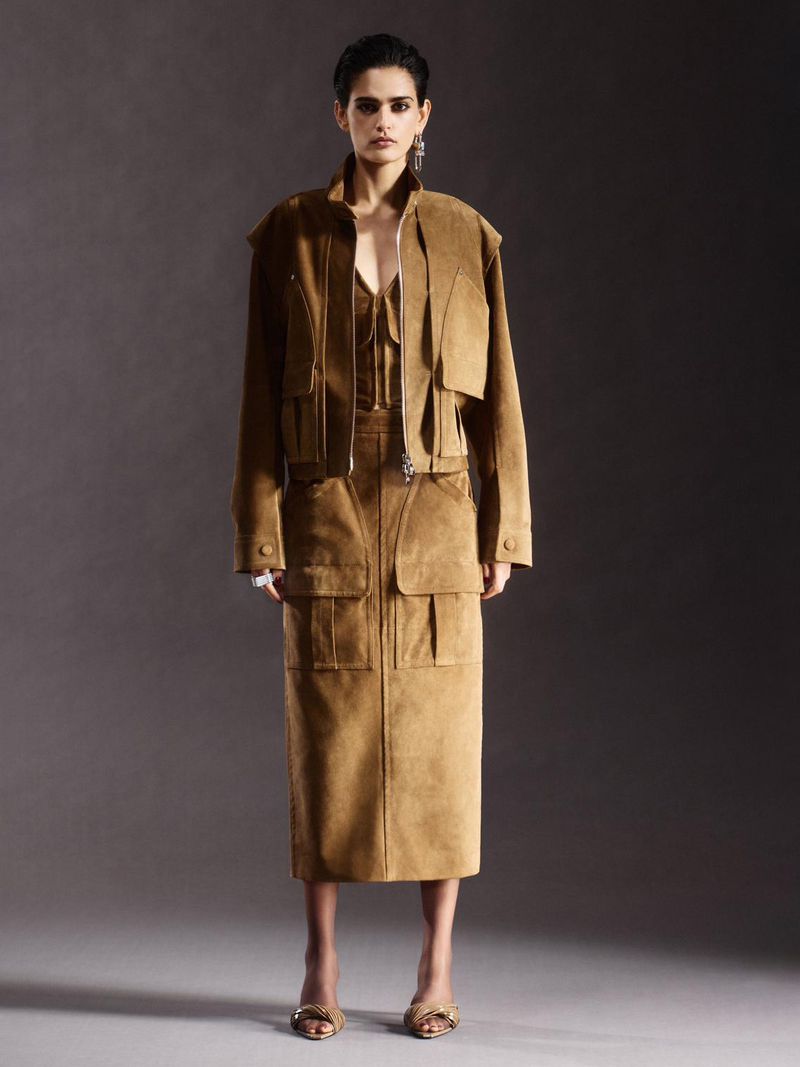 A woman in a tan jacket and matching skirt poses against a dark gray background, showcasing an editorial fashion look with soft lighting.