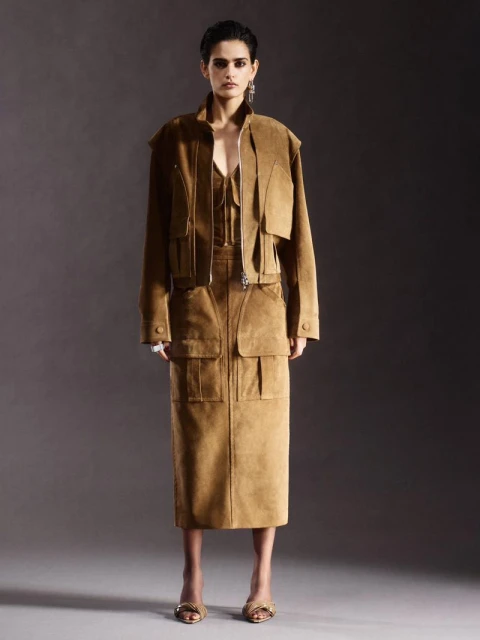 A woman in a tan jacket and matching skirt poses against a dark gray background, showcasing an editorial fashion look with soft lighting.