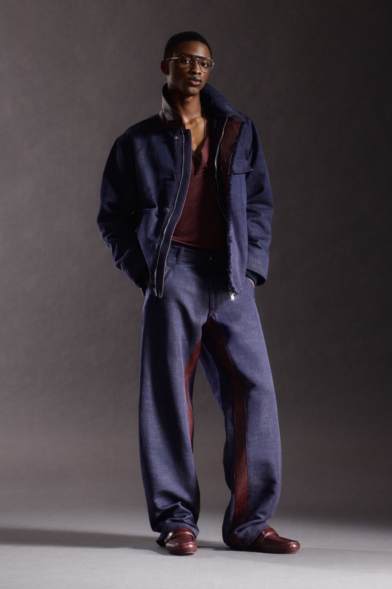 Full-body studio shot of a black male model in baggy denim jacket and pants, with burgundy shirt and leather shoes, against gray backdrop.