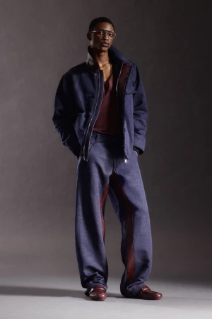 Full-body studio shot of a black male model in baggy denim jacket and pants, with burgundy shirt and leather shoes, against gray backdrop.