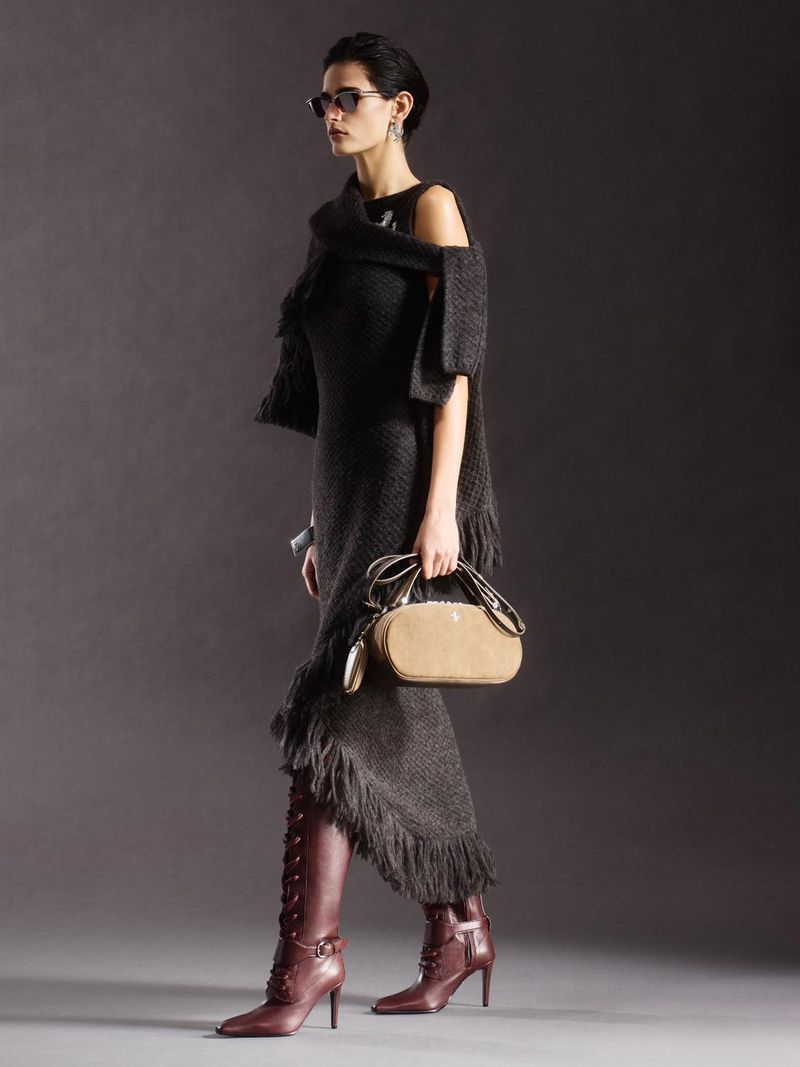 A woman models a dark, fringed dress and matching shawl with sunglasses and heeled boots in a full-length studio shot on gray.