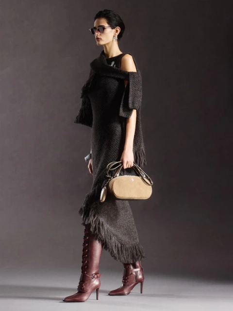 A woman models a dark, fringed dress and matching shawl with sunglasses and heeled boots in a full-length studio shot on gray.
