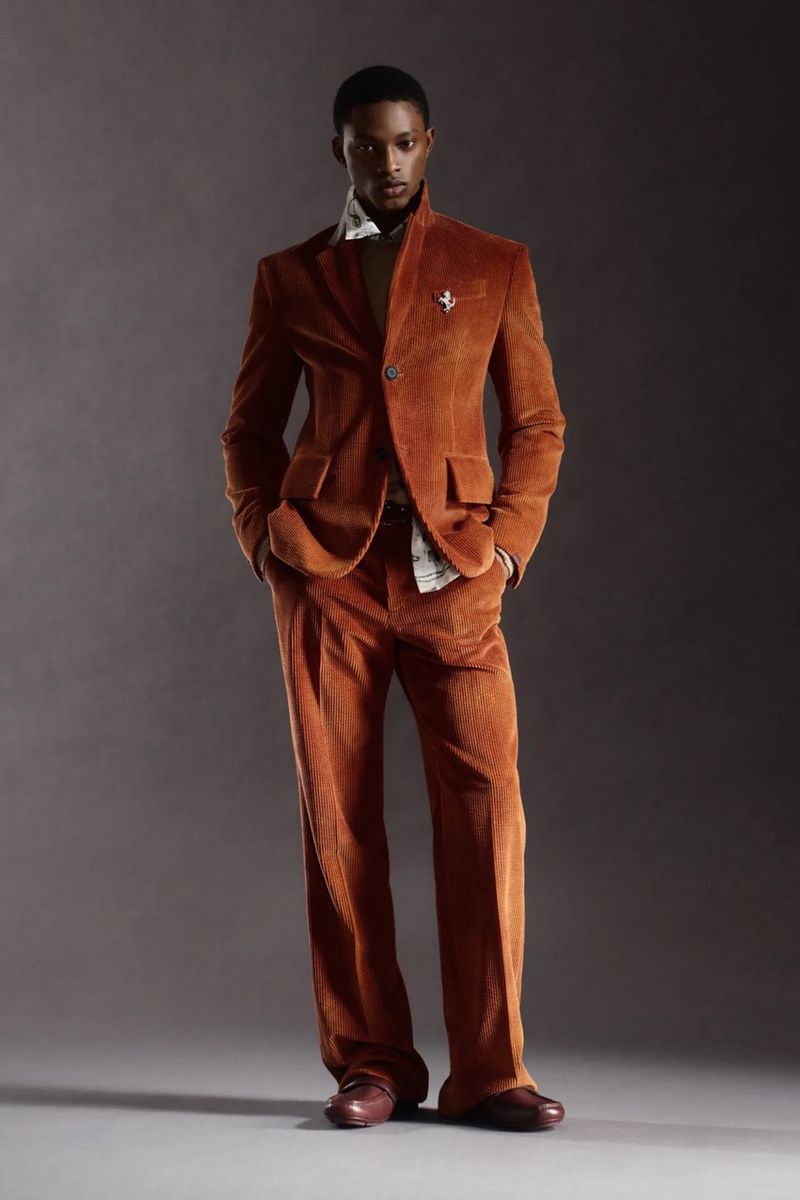 Full-length studio shot of a Black male model in an orange corduroy suit, with a beige shirt and brown leather shoes against a gray backdrop.