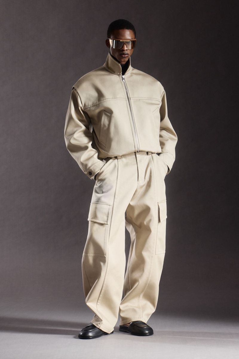 Fashionable man in beige jacket and cargo pants, accessorized with modern glasses and black loafers, posing in a studio setting.
