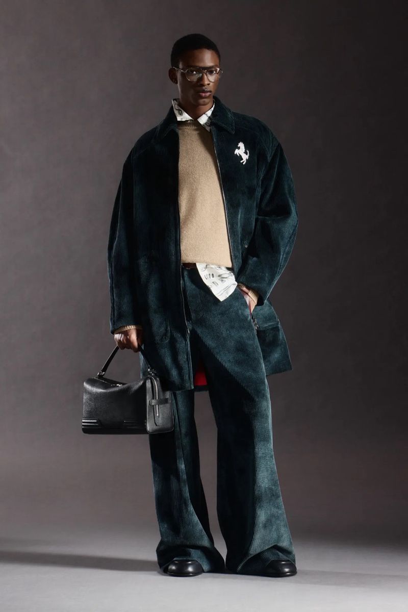 A black male model poses in a studio wearing a dark green corduroy overcoat and pants, a tan crew neck sweater, and carrying a black bag.