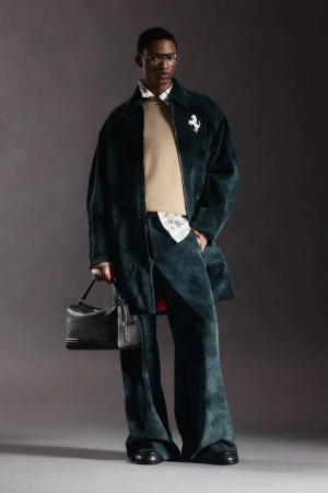A black male model poses in a studio wearing a dark green corduroy overcoat and pants, a tan crew neck sweater, and carrying a black bag.