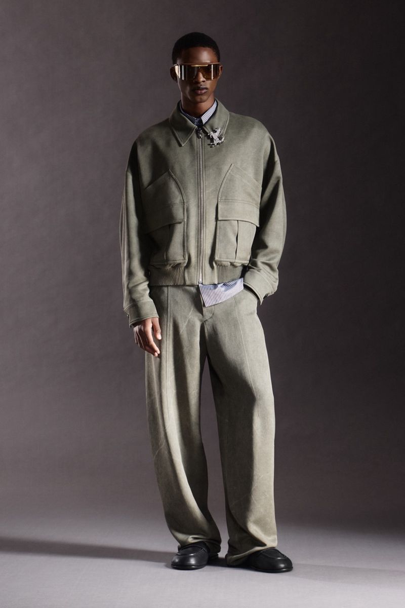 Fashionable adult male model poses in a studio wearing a matching light green jacket, trousers, black shoes, and gold tinted glasses.