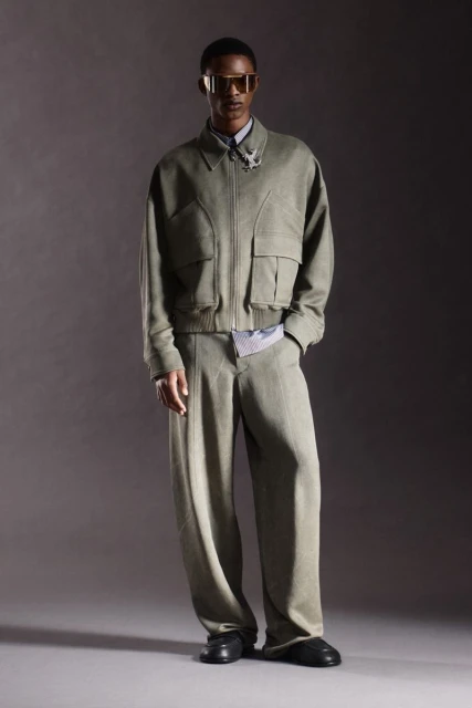 Fashionable adult male model poses in a studio wearing a matching light green jacket, trousers, black shoes, and gold tinted glasses.