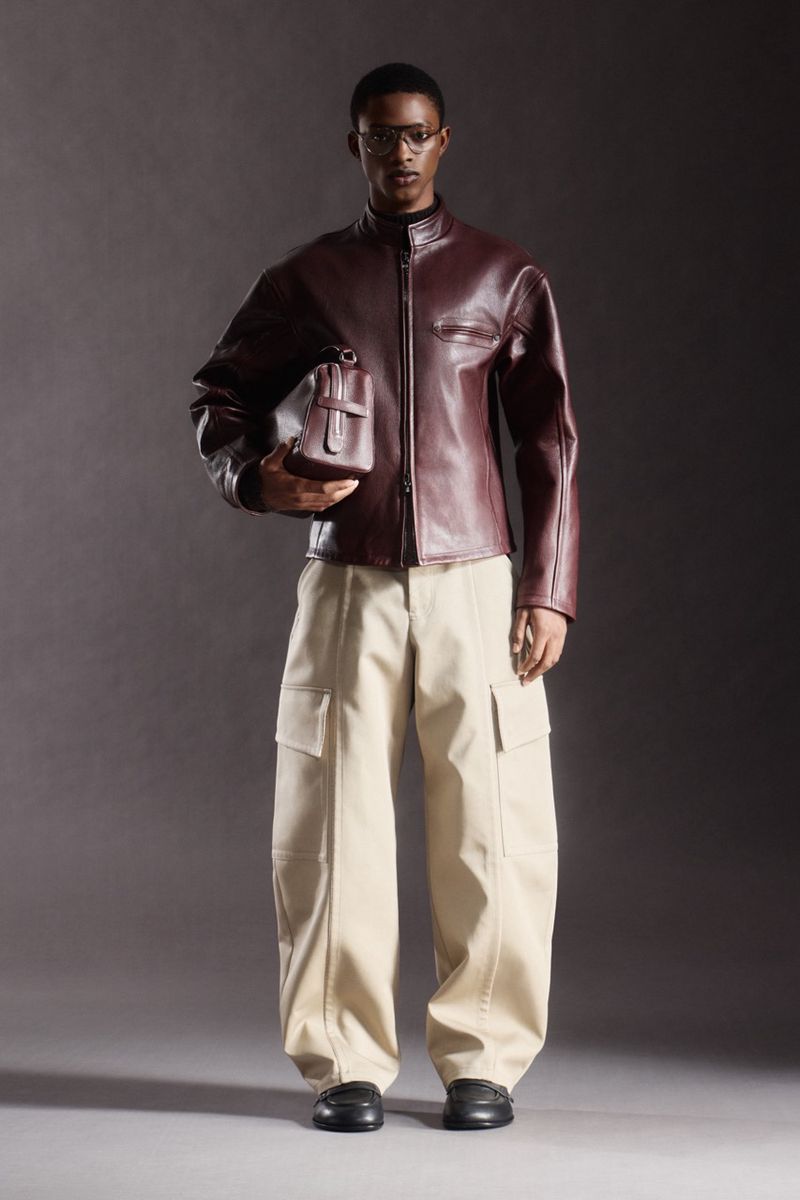 Male model poses in studio wearing burgundy leather jacket, khaki cargo pants, black shoes, and glasses, holding burgundy bag.