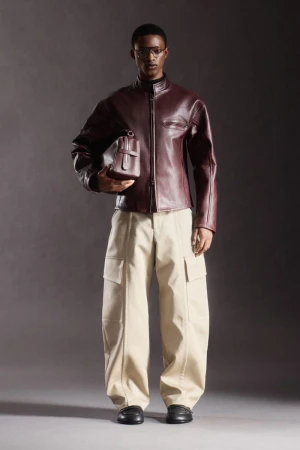 Male model poses in studio wearing burgundy leather jacket, khaki cargo pants, black shoes, and glasses, holding burgundy bag.