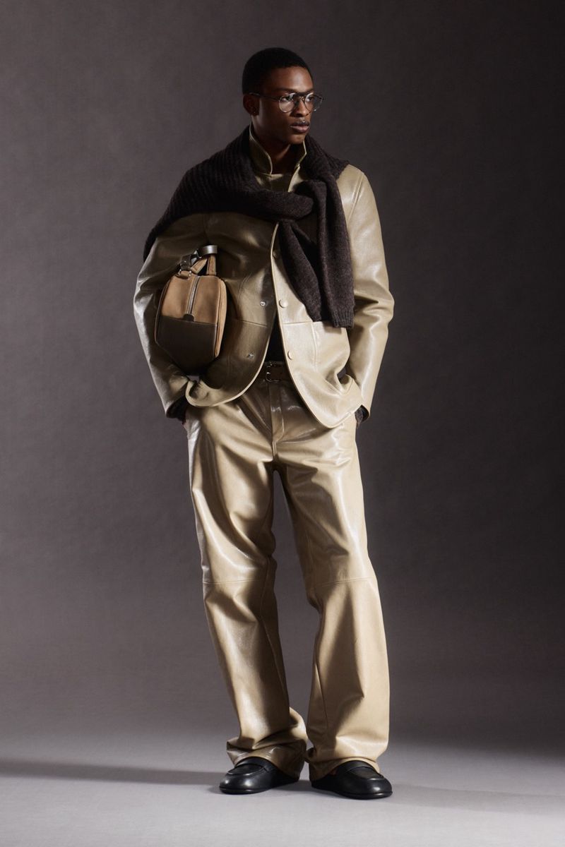 Man in beige suit and dark scarf, posing in a studio. He is holding a beige handbag and wearing glasses, with a neutral facial expression.