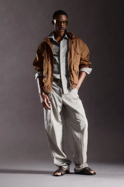 Adult male in a grey button-down shirt, brown jacket, and light-colored pants poses in sandals and futuristic sunglasses in a studio.