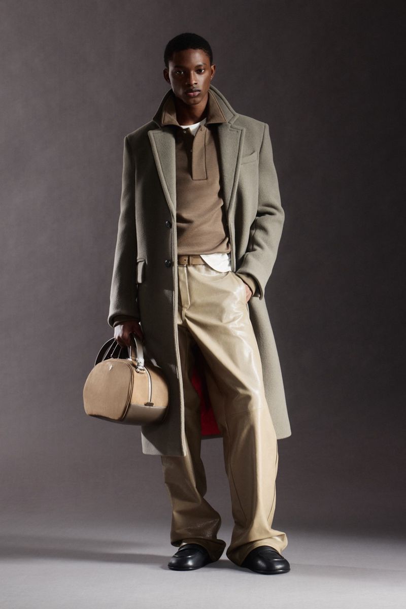 A dark-skinned male model poses indoors wearing a coat, polo shirt, and pants, carrying a tan leather bag, against a grey backdrop.