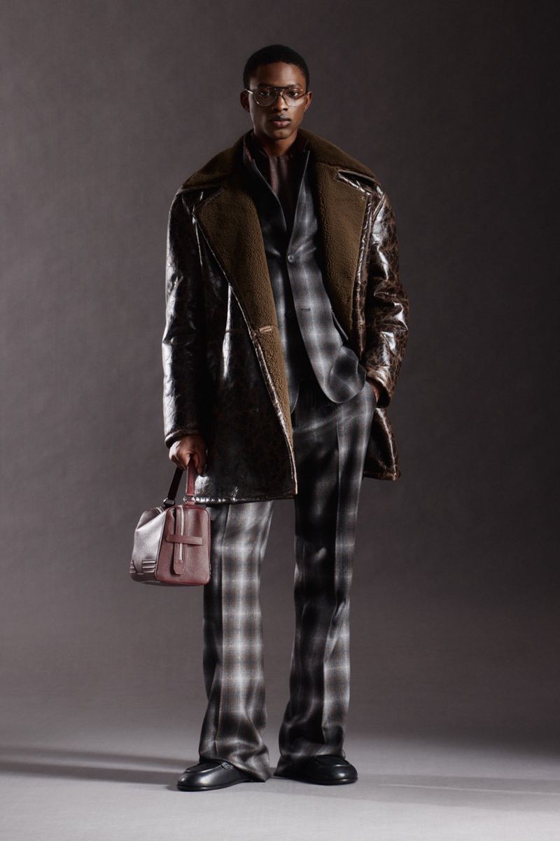 Full-shot of a man in a patterned suit and a leather coat, holding a purse. The studio backdrop is a gradient of muted brown.
