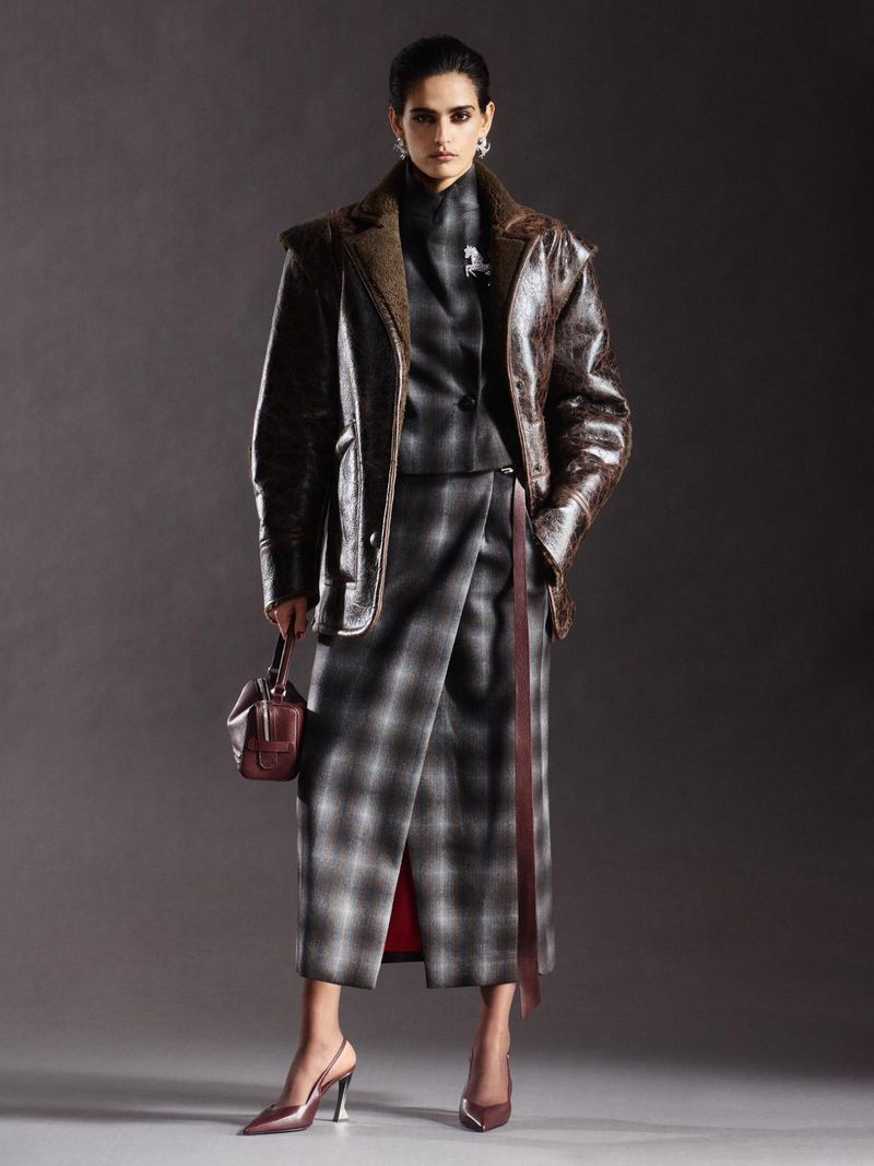 Fashionable woman in studio wearing a plaid skirt suit with a brown leather jacket, holding a small purse, and wearing heels.