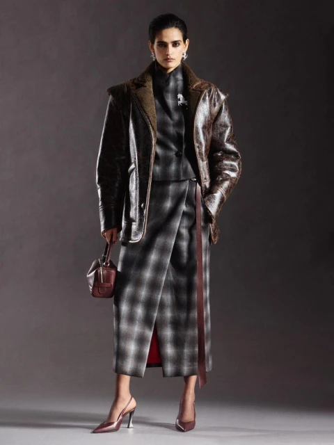 Fashionable woman in studio wearing a plaid skirt suit with a brown leather jacket, holding a small purse, and wearing heels.