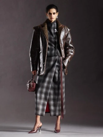 Fashionable woman in studio wearing a plaid skirt suit with a brown leather jacket, holding a small purse, and wearing heels.