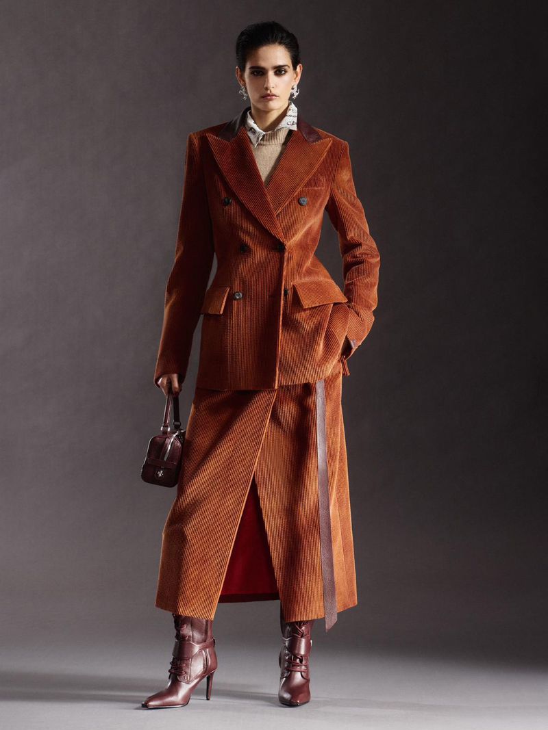 A woman in a corduroy suit and matching boots poses in a studio setting holding a small handbag, showcasing a high-fashion fall look.