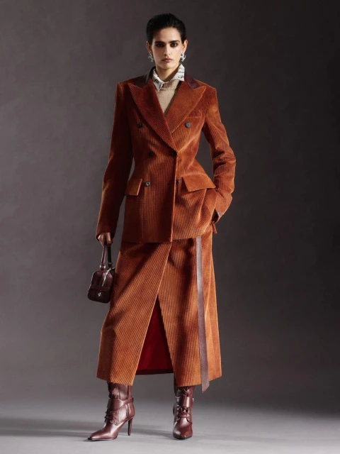 A woman in a corduroy suit and matching boots poses in a studio setting holding a small handbag, showcasing a high-fashion fall look.