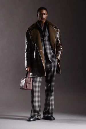Model Nonso Ojukwu in a shearling-lined leather coat and plaid suit holding a burgundy bag against a grey studio backdrop.