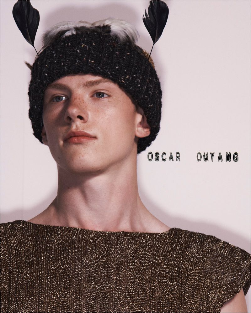 Close-up studio shot of a young adult man with light skin wearing a decorative hat and a textured top against a plain backdrop.