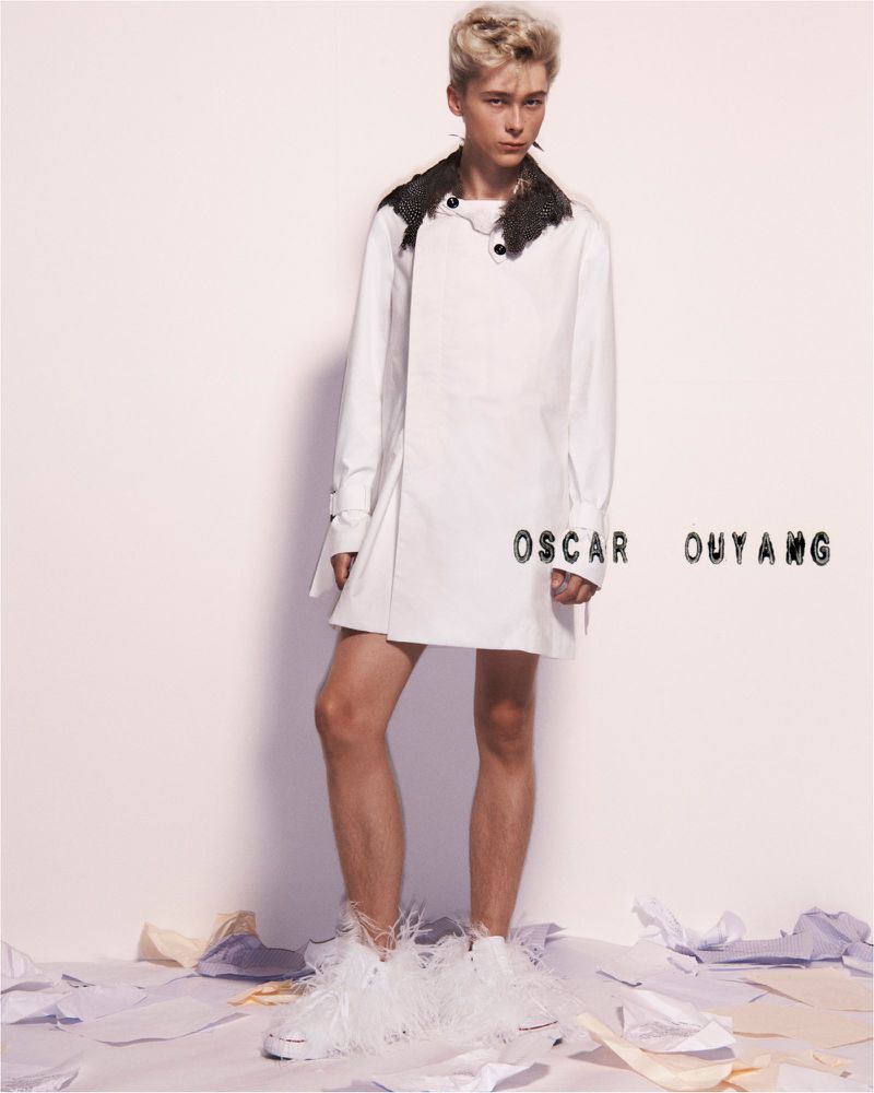 Full studio shot of a young adult in a white coat with fur collar and feathered shoes, standing on a white floor with scattered papers.
