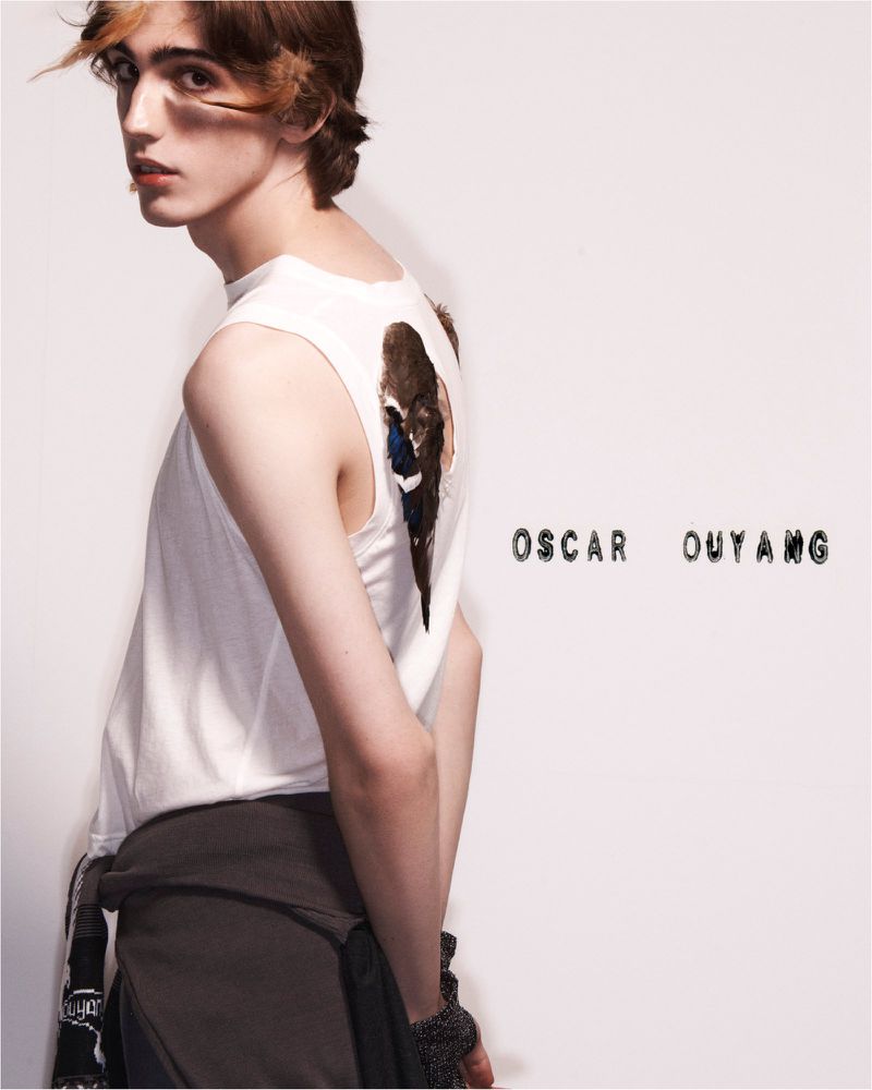 Studio shot of a young male adult posing in a white top with graphic detail, accessorized with a grey skirt and bracelets against a white backdrop.