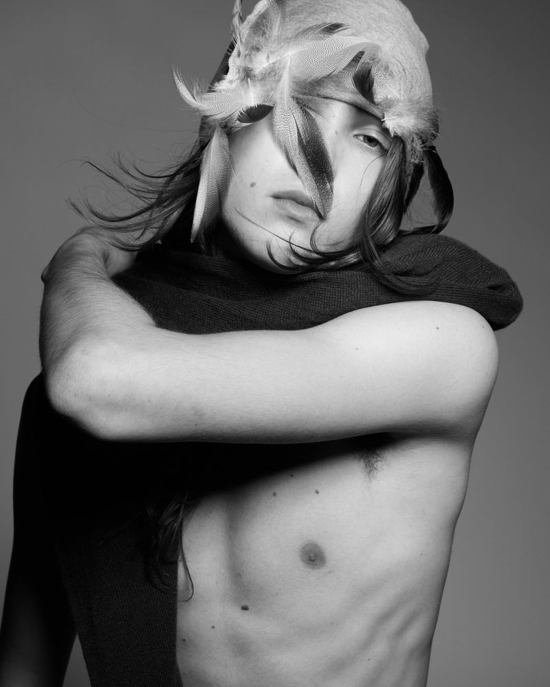 Black and white portrait of a shirtless adult male wearing a feather headpiece looking toward the camera in a photo studio.