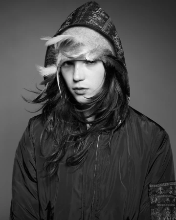Monochrome portrait of a young adult with long hair wearing a hooded jacket and feathered headpiece against a solid gray background.