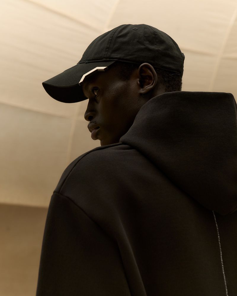 Profile of a black male adult wearing a black baseball cap and black hooded sweatshirt against a soft tan neutral backdrop.