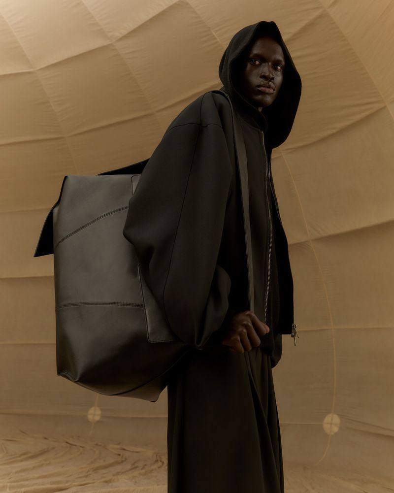 Fashion shot of a black male model wearing a hooded jacket, pants, and carrying a large leather bag in a neutral-toned indoor environment.
