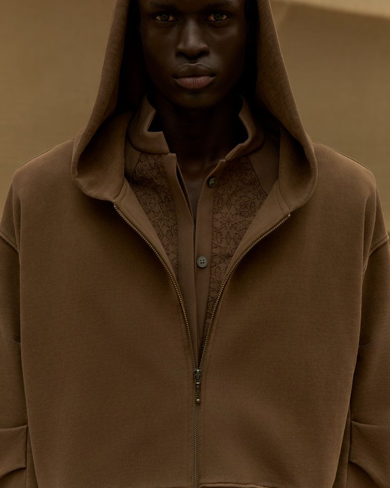 A dark-skinned adult male with a serious expression poses in a brown hooded sweatshirt, worn over a button-down shirt, against a neutral background.
