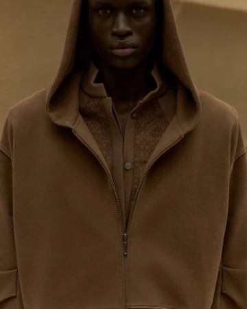 A dark-skinned adult male with a serious expression poses in a brown hooded sweatshirt, worn over a button-down shirt, against a neutral background.