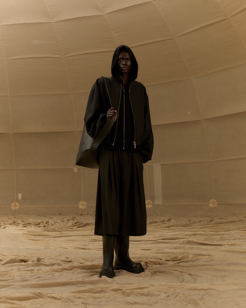 Full-length portrait of a Black male wearing a black hooded sweatshirt, long black skirt, and boots, standing in a domed structure.