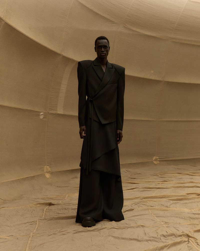 A slender Black male model wearing a dark green draped suit stands against a beige fabric backdrop in a fashion editorial.