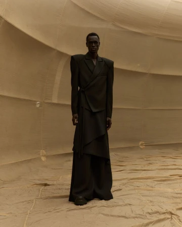 A slender Black male model wearing a dark green draped suit stands against a beige fabric backdrop in a fashion editorial.