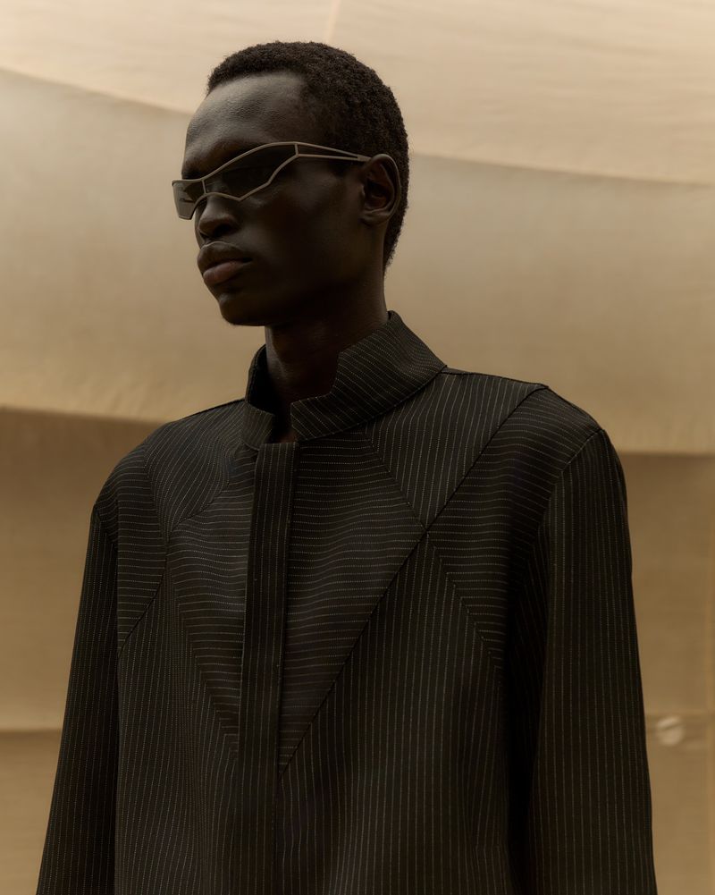 Model Daniel Ajang wearing futuristic wireframe sunglasses and a dark pinstripe coat against a warm neutral studio backdrop.