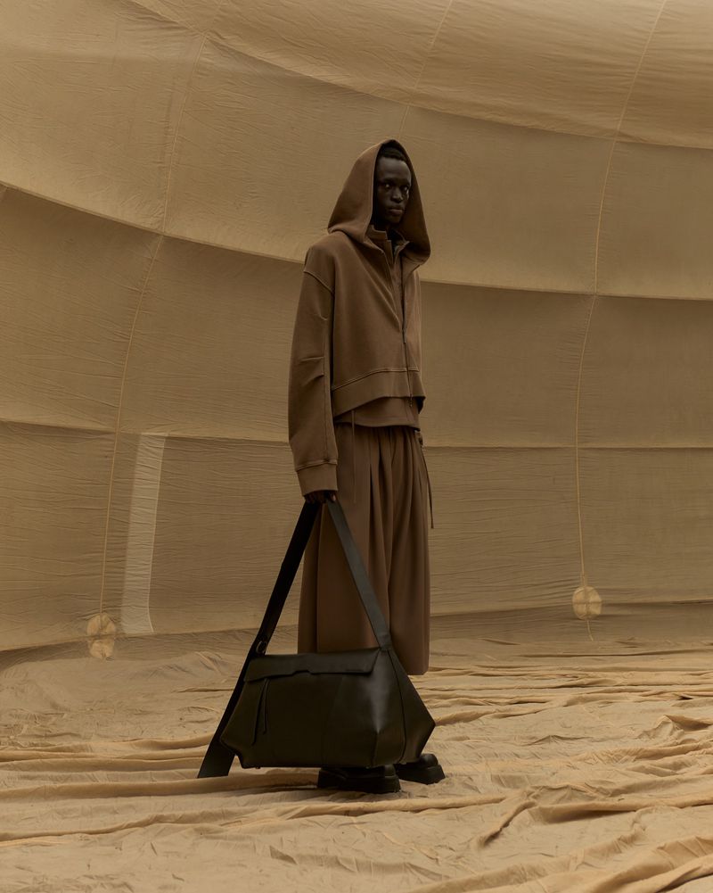 A person with dark skin and a hooded brown outfit carrying a large black bag, posed in a tan-colored, dome-like architectural space.