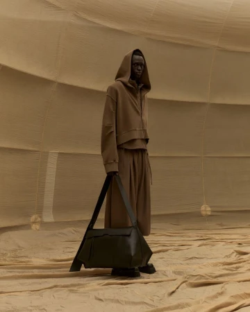 A person with dark skin and a hooded brown outfit carrying a large black bag, posed in a tan-colored, dome-like architectural space.