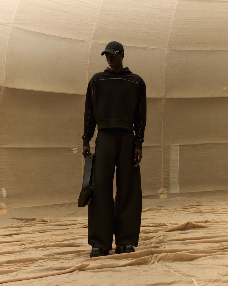 Dark-skinned male model in oversized black pants, hoodie, and cap stands in large spherical room with natural light. Neutral background.