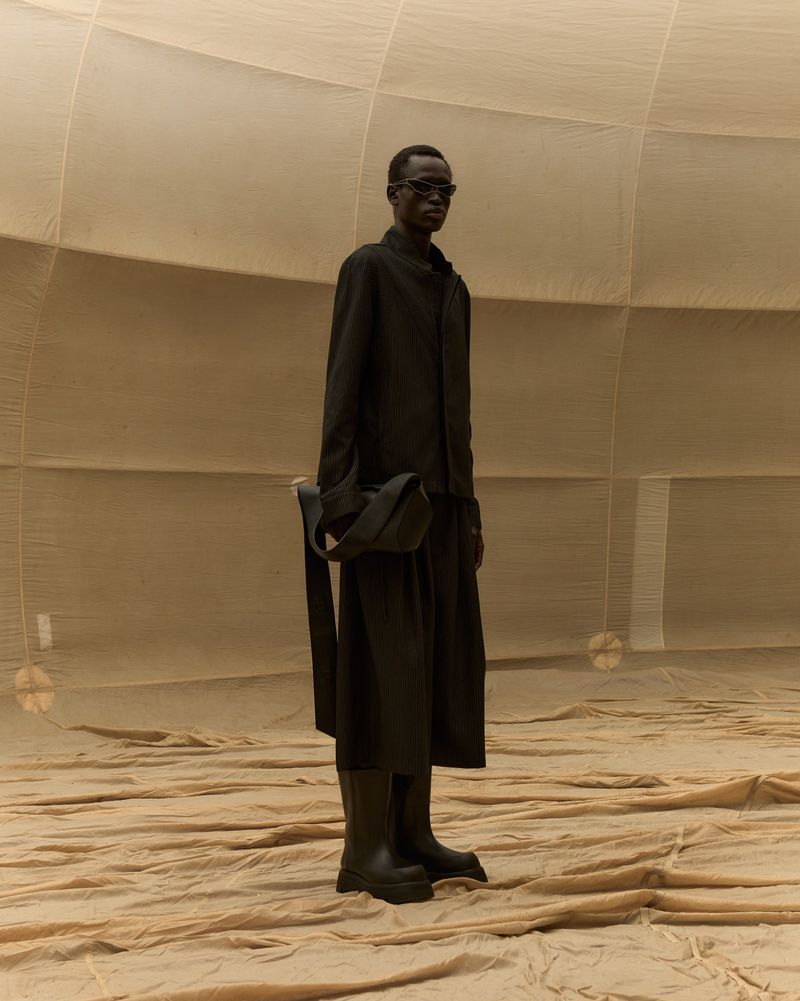 Fashionable adult male poses in a monochrome black outfit with boots, glasses and bag in a textured, earth-toned setting.