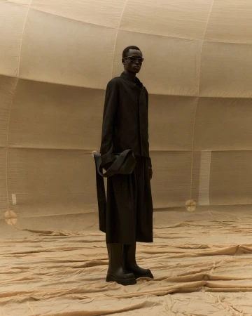 Fashionable adult male poses in a monochrome black outfit with boots, glasses and bag in a textured, earth-toned setting.
