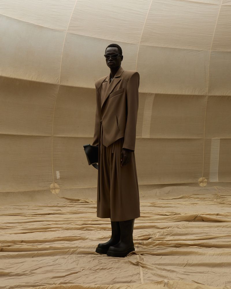 Fashion model in a tan suit, black boots, and sunglasses stands in a photo studio with tan backdrops in a full body shot.