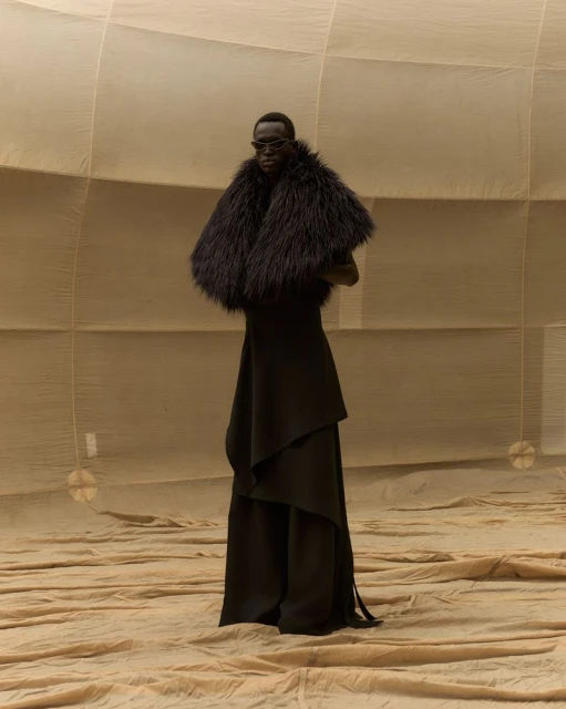 A black female model wearing sunglasses and a black fur stole poses in a tan fabric environment with a dramatic draped dress.