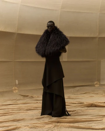 A black female model wearing sunglasses and a black fur stole poses in a tan fabric environment with a dramatic draped dress.