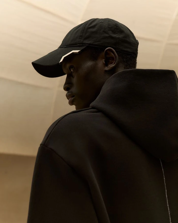 Black man wearing a black baseball cap and a black hooded sweatshirt viewed from behind looking sideways in warm lighting.