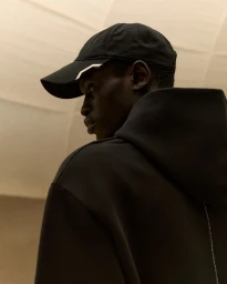 Black man wearing a black baseball cap and a black hooded sweatshirt viewed from behind looking sideways in warm lighting.